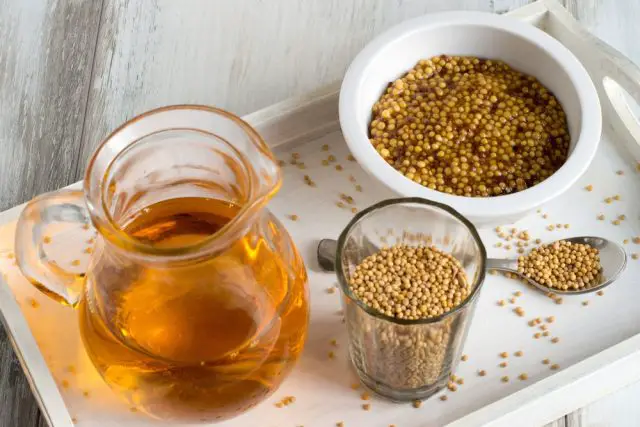 A cup with mustard, a glass with mustard seeds and a jug with mustard oil on a white wooden tray.