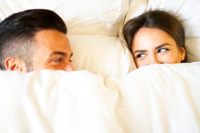 Boyfriend and girlfriend laying in a bed with white sheets - Young and beautiful couple happily looking at each other during bed time