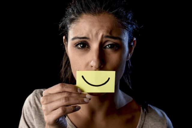 Young beautiful Latin sad and depressed latin girl holding paper hiding her mouth behind a fake drawn smile pretending to be happy in depression 