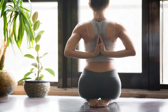 Young woman practicing yoga with namaste behind the back, sitting in seiza exercise, vajrasana pose, working out